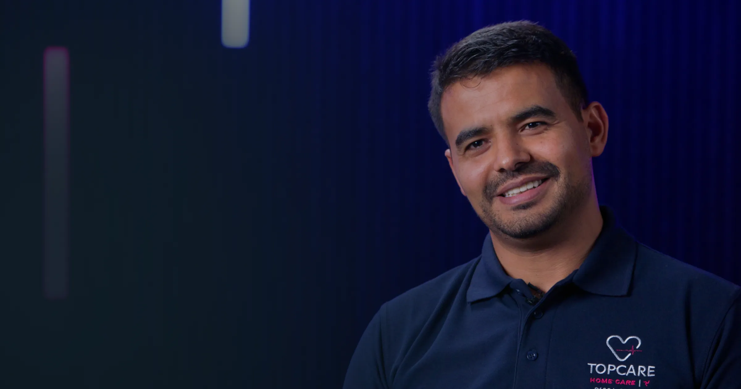 A man in a navy polo with a heart logo smiles warmly against a dark, blurred background. Soft lighting gives a calm and professional atmosphere.